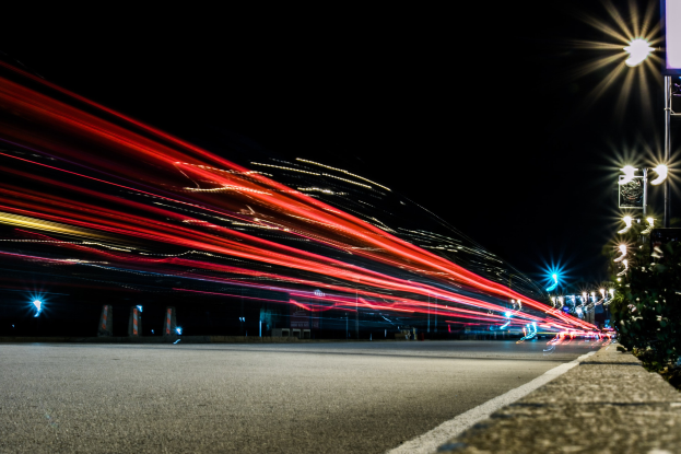 Langzeitaufnahme einer nächtlichen Stadtstraße mit beleuchtetem Verkehr und Straßenlaternen, die Lichtspuren gegen einen dunklen Hintergrund werfen, gesäumt von Pflanzen auf der rechten Seite.