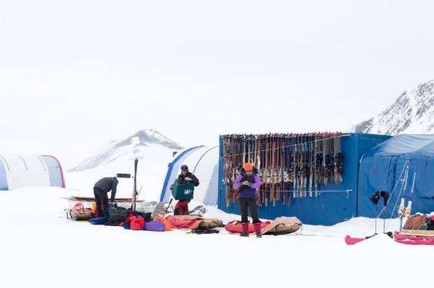 Drei Personen stehen auf einer schneebedeckten Landschaft mit verstreuten Taschen, Zelten mit Skiern darauf und schneebedeckten Hügeln im Hintergrund unter einem klaren Himmel.