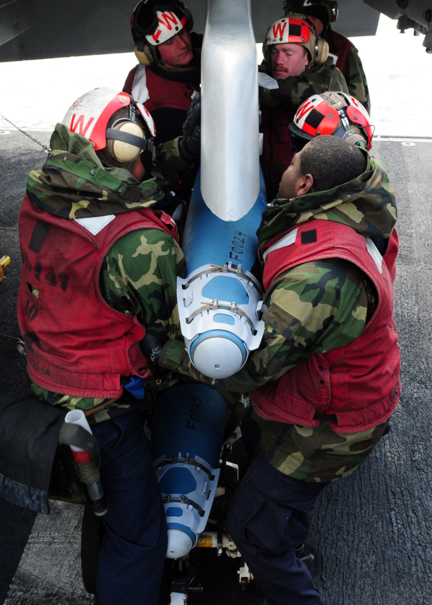Eine Gruppe von Männern in roten Jacken und Helmen steht um eine Rakete herum, einer hält ein Objekt, und im Hintergrund ist ein Flugzeug zu sehen.