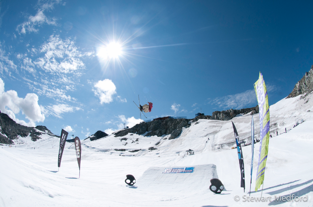 Eine Person in Skiausrüstung schwebt in der Luft mit schneebedeckten Bergen im Hintergrund.