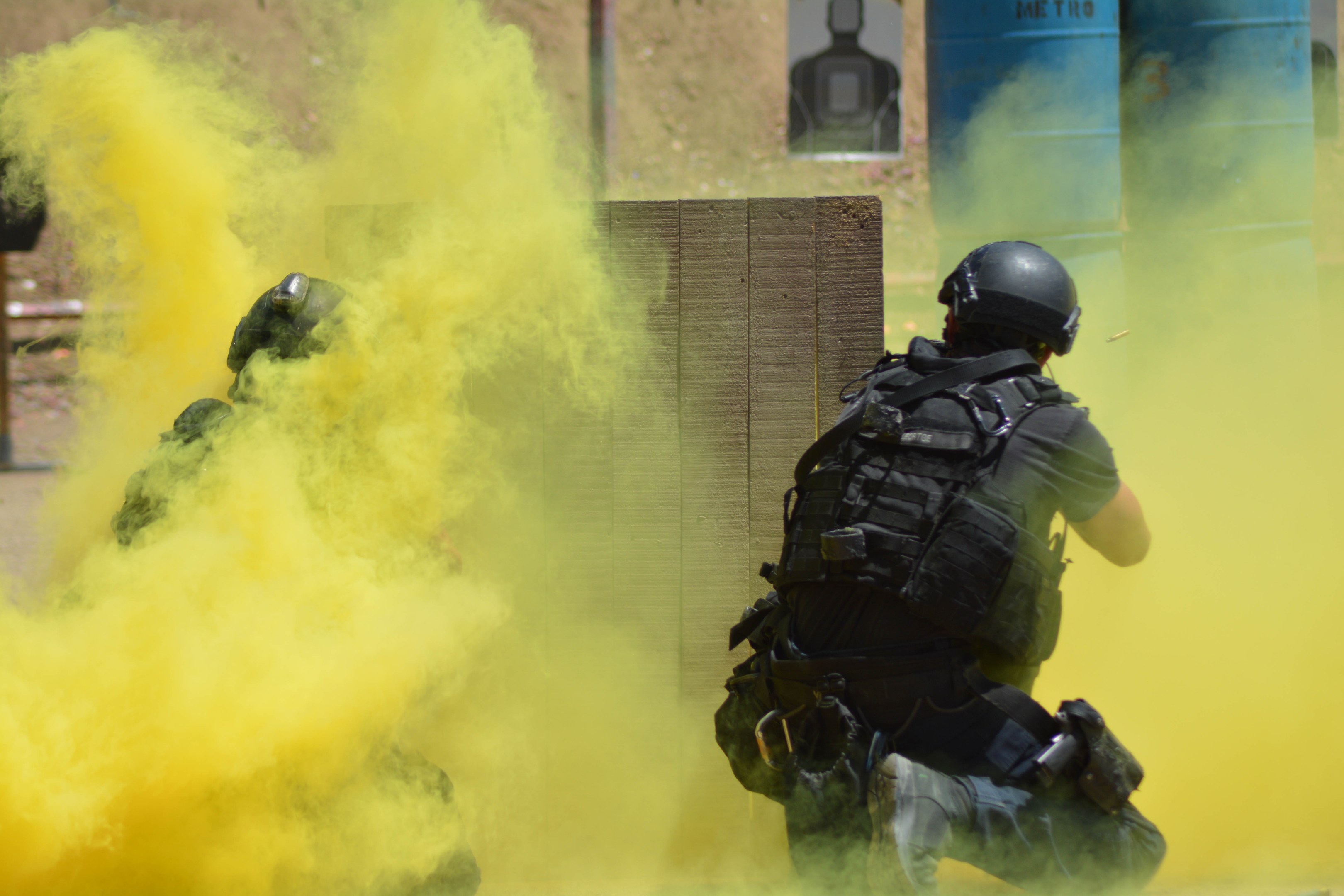 Zwei Polizisten in Einsatzausrüstung mit gelbem Rauch aus dem Mund, die neben einer Wand mit Text und anderen Gegenständen im Hintergrund stehen.