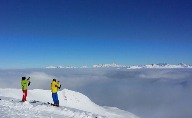 Zwei Personen stehen auf Snowboards in einer verschneiten Landschaft, halten Gegenstände und haben Hügel und Himmel im Hintergrund.