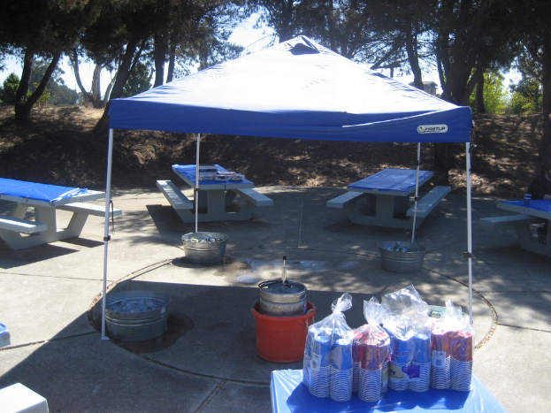 Partybecher in Verpackungen mit Preisschildern auf einem Tisch, Eimer mit Gegenständen unter einem Zelt, Bänke mit Gegenständen um das Zelt, Bäume und ein hölzernes Haus im Hintergrund.