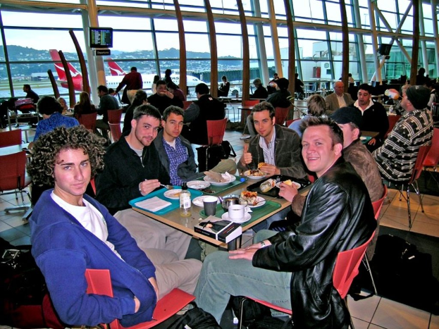 Eine Gruppe von Menschen sitzt um einen Tisch mit roten Stühlen in einem Flughafen, isst und spricht, mit einer Glaswand, einem Hügel, einem Flugzeug und einem Fernseher im Hintergrund.