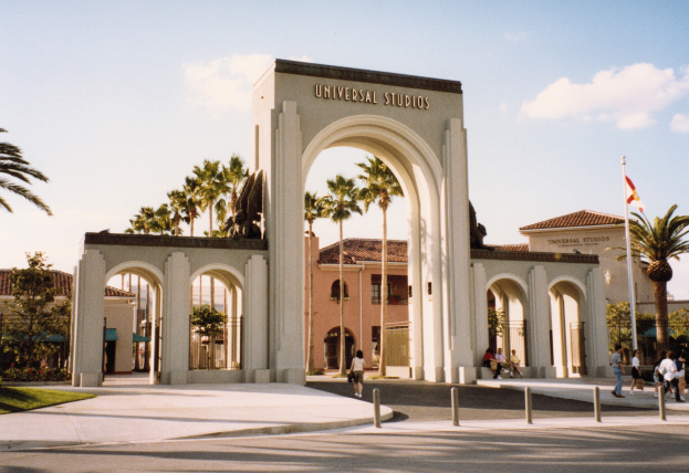 Eingang zum Universal Studios Hollywood mit einem Bogen, einer Statue, Menschen, Bäumen, Gebäuden, einem Fahnenmast mit einer Fahne und einem bewölkten Himmel.