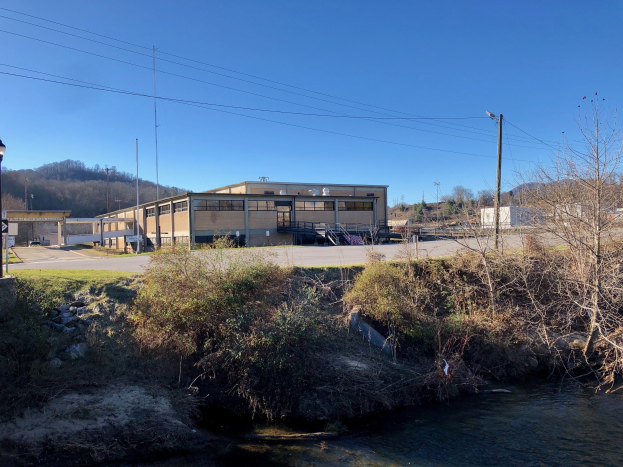 Ein Fluss fließt durch eine grüne Wiese neben einem Gebäude neben Bäumen, Pflanzen, Gras, Steinen, Straßenlaternen, Strommasten, Kabeln, Hügeln und einem blauen Himmel, wobei das Gebäude als Standort einer geplanten Wasseraufbereitungsanlage vorgeschlagen wird.