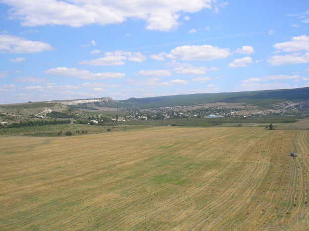 Ein Traktor auf einem kultivierten Feld umgeben von Bäumen, Gebäuden, Hügeln und einem bewölkten Himmel.