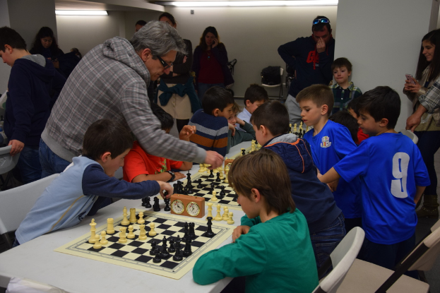 Kinder spielen Schach bei einem Wettbewerb, während Erwachsene im Hintergrund und am Rand zuschauen.