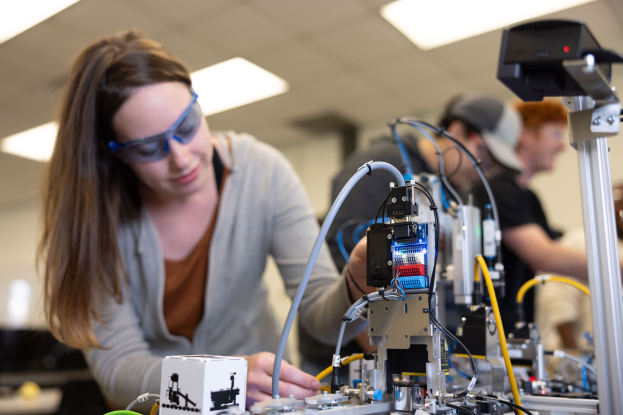 Eine Frau mit Schutzbrille arbeitet an einem Roboter in einem Labor umgeben von anderen, mit Werkzeugen und Komponenten auf dem Tisch unter Deckenbeleuchtung.