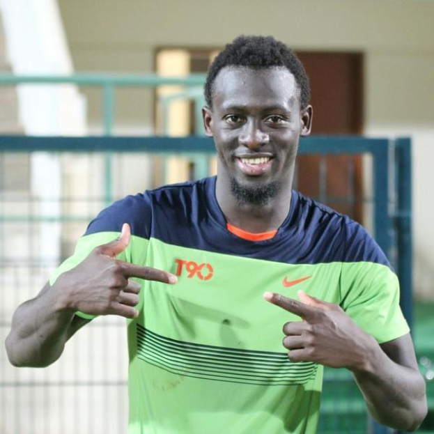 Ein Mann in einem grünen und blauen Hemd zeigt mit dem Finger auf die Kamera, mit einem unscharfen Hintergrund aus einem Geländer, einer Wand und einer Tür.