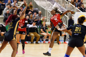Eine Gruppe von Frauen beim Volleyballspielen auf einem Platz mit einem Ball in der Mitte, während Zuschauer auf Stühlen und Geländern im Hintergrund sitzen und Flaschen auf dem Boden liegen.