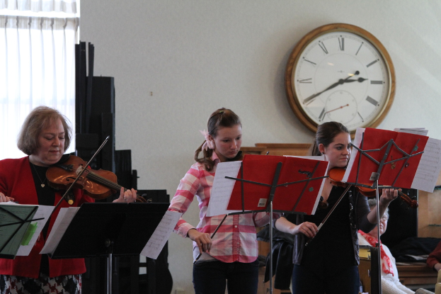 Drei Frauen auf der Bühne mit Violinen stehend und spielend, mit Notenpulten und Noten davor, während ein Zuschauer im Vordergrund sitzt; eine Uhr ist an der weißen Wand im Hintergrund angebracht, und ein weißer Vorhang ist links im Bild zu sehen.