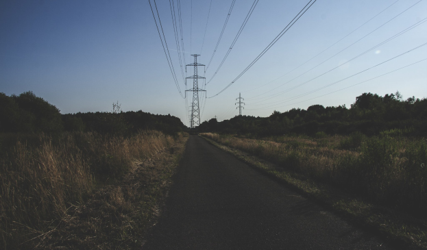 Straße mit Überlandleitungen, gesäumt von grünem Gras und Bäumen, führt zu entfernten Übertragungsmasten.