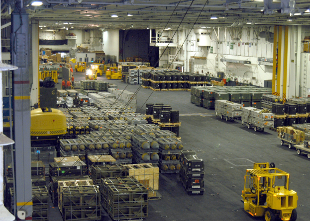 Ein großes Lagerinnere voller Numerous Trucks, Container, Menschen, Lichter, Stangen und andere Gegenstände mit einem prominenten Rad.