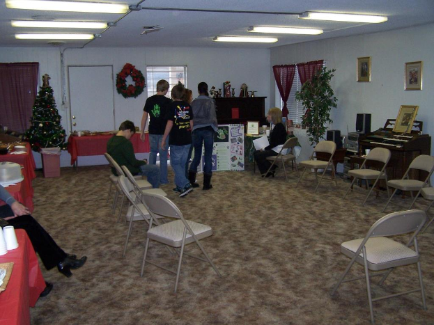 Ein Raum mit mehreren Stühlen, Tischen und Menschen, einige stehen, andere sitzen. Auf den Tischen befinden sich Tassen und Gefäße. Im Hintergrund eine Wand mit einem Strauß, Vorhang, Tür, Fotorahmen und Fenstern. Ein Weihnachtsbaum und ein Piano auf der rechten Seite. Leuchtstoffröhren an der Decke.