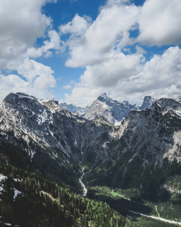 Atemberaubender Ausblick auf ein Gebirgsmassiv von einem Hügel aus, mit grünen Bäumen und schneebedeckten Gipfeln unter einem Himmel voller weißer Wolken.
