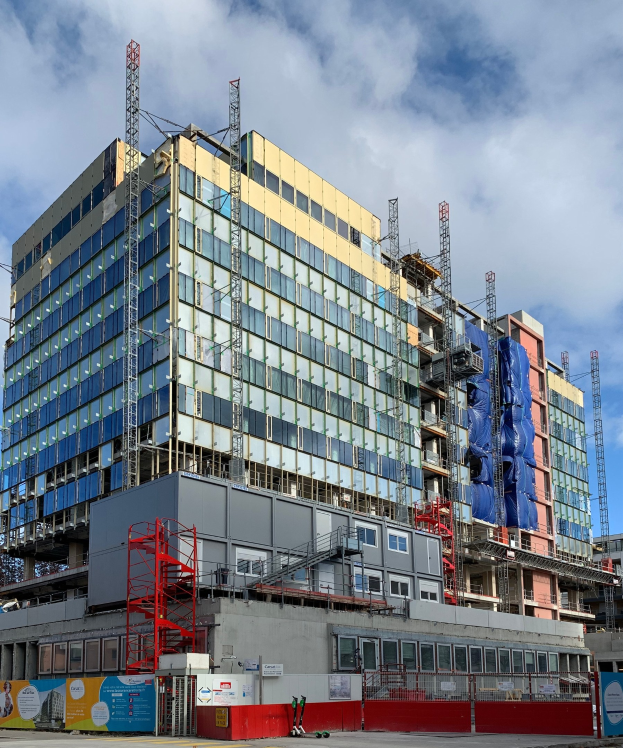 Großes Gebäude im Bau in einer Stadt mit Glasfenstern, Metallrahmen, Treppe, Umzäunung, Schild, Versorgungsmasten, Straßenlaterne, nahen Bäumen und einem bewölkten Himmel.