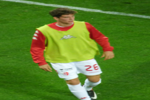 Ein Mann in einem gelben und roten T-Shirt läuft auf einem Fußballfeld, das mit weißen Linien markiert ist und trägt Fußballschuhe.