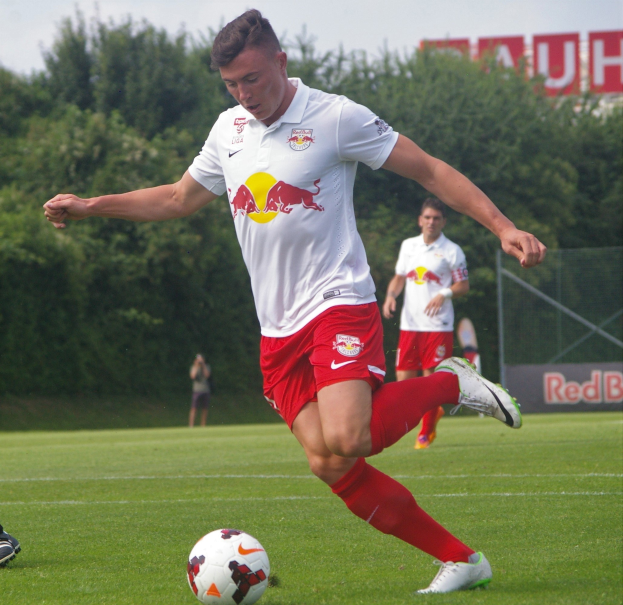 Ein professioneller Fußballspieler in einem roten und weißen Trikot kickt einen Fußball auf einem Rasenfeld mit Bäumen und einem klaren blauen Himmel im Hintergrund, wobei ein Schild mit der Aufschrift "RB Leipzig" sichtbar ist.
