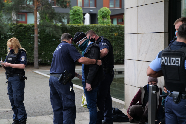 Gruppe von Polizeibeamten vor einem Gebäude mit einer Frau, die ein Buch und einen Stift hält, umgeben von sitzenden und stehenden Personen, mit Bäumen und Gebäuden im Hintergrund.