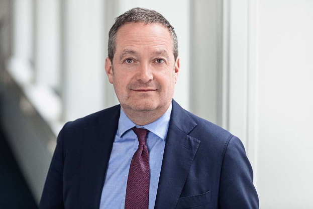 Ein Mann im Anzug und Krawatte steht vor einem Fenster in einer professionellen Umgebung und scheint der neu ernannte CEO des Unternehmens zu sein.