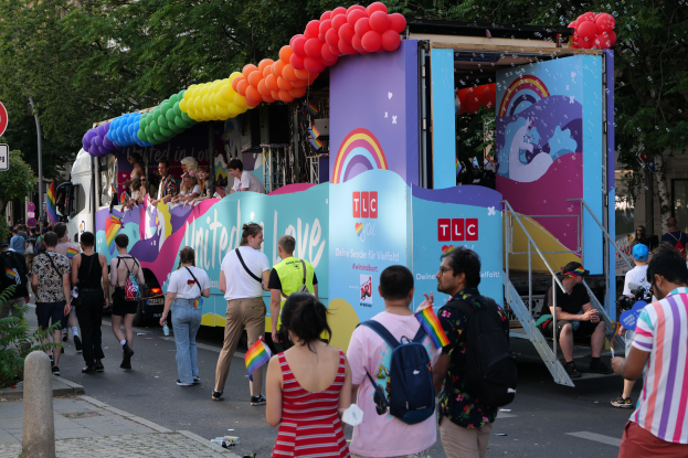 Eine Gruppe von Menschen geht auf einer Straße neben einem Lastwagen mit bunten Luftballons, mit Schildern an der Straße und Bäumen und Gebäuden im Hintergrund, was auf eine Pride-Parade in Paris hindeutet.