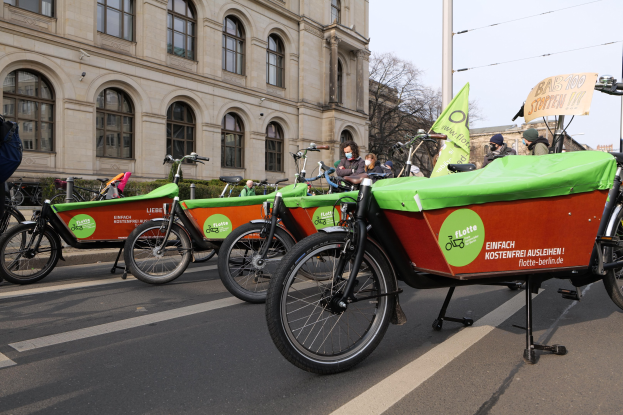 Eine Gruppe von Fahrrädern, die auf einer Straße mit einer Person in der Nähe geparkt sind, Gebäude, Bäume, Pfosten, Drähte und einen klaren blauen Himmel im Hintergrund und ein Banner im Vordergrund, das eine Fahrrad-Sharing-Kampagne ankündigt.