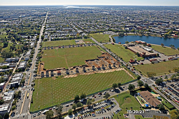 Eine Luftaufnahme einer Stadt mit Gebäuden, Bäumen, Straßen mit Fahrzeugen, Wasser und einem bewölkten Himmel, die ein großes zentrales Feld zeigt, das für einen Park vorgesehen ist.