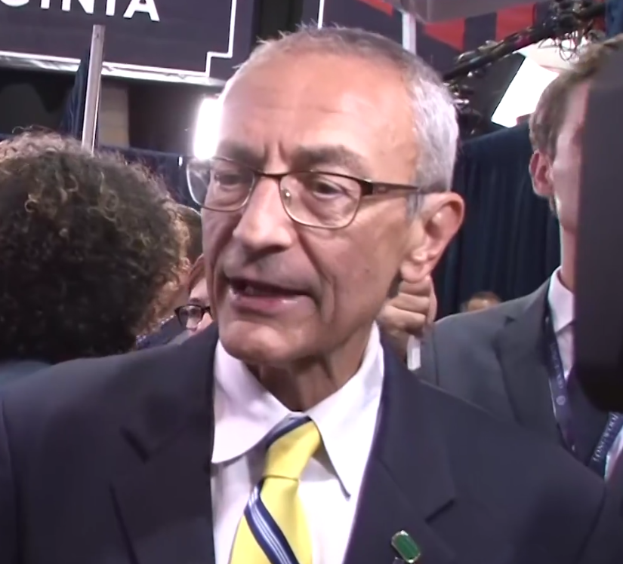 Ein Mann im Anzug und mit Krawatte, der eine Brille trägt, spricht zu Reportern mit einem Mikrofonständer vor sich, umgeben von einer Gruppe von Menschen, mit Lampen und einer Tafel im Hintergrund.