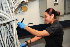 Eine Frau in einem schwarzen Hemd und blauen Handschuhen arbeitet an einem Computer, hält ein Objekt und hat zahlreiche Kabel links von sich und eine wandmontierte Box im Hintergrund, was den Erfolg eines Frauen-Ingenieurprogramms symbolisiert, das einen renommierten Preis gewonnen hat.