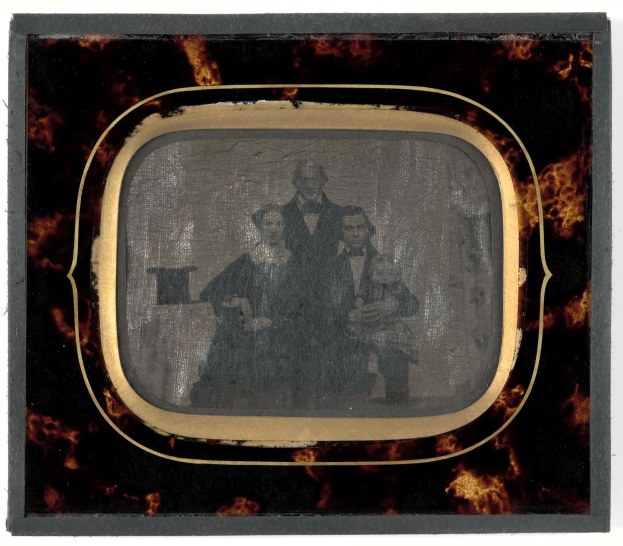 Ein altes Foto eines Mannes in Anzug und Krawatte und einer Frau in unbekannter Kleidung, die vor einem Fernseher stehen und ernst schauen.