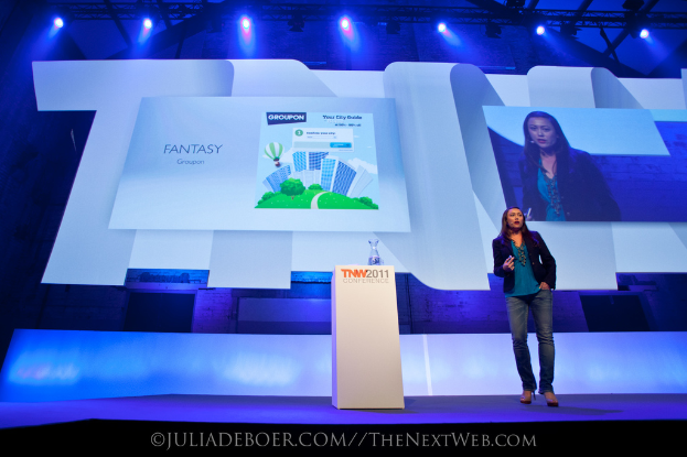 Eine Frau steht an einem Rednerpult auf einem Podium, mit einem Bildschirm und Lampen im Hintergrund.