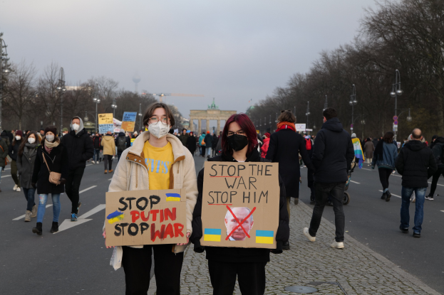 Eine Gruppe von Menschen mit Masken und Schildern, auf denen "Stop the War" steht, geht eine von Bäumen gesäumte Straße in Berlin, Deutschland, entlang, wobei im Hintergrund ein Tor zu sehen ist.