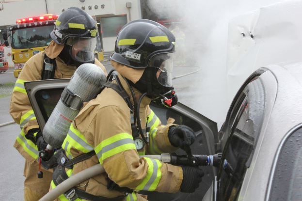 Zwei Feuerwehrleute in Schutzausrüstung verwenden einen Schlauch, um ein brennendes Auto zu löschen, während im Hintergrund Fahrzeuge und ein Gebäude zu sehen sind.