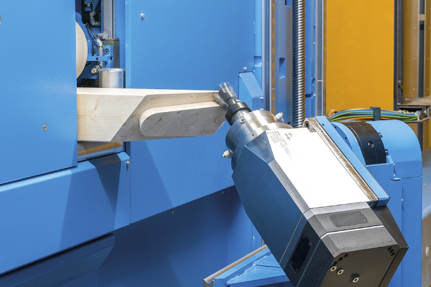 Eine CNC-Fräsmaschine schneidet ein Holzstück in einer Fabrikumgebung, umgeben von Kabeln und anderen Gegenständen, mit einer Wand im Hintergrund.