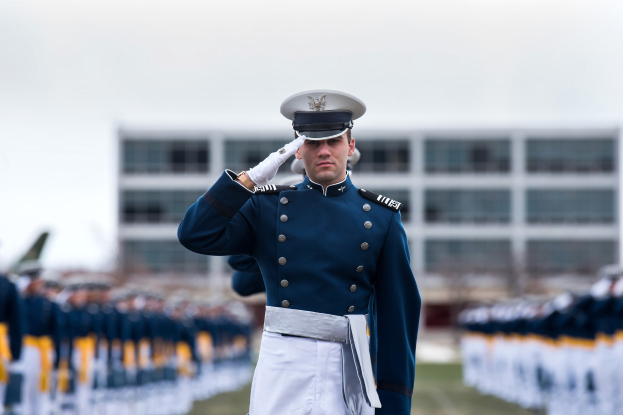 Ein Mann in militärischer Uniform salutiert auf einer Abschlussfeier umgeben von einer Gruppe von Menschen, mit einem Gebäude und Himmel im Hintergrund.