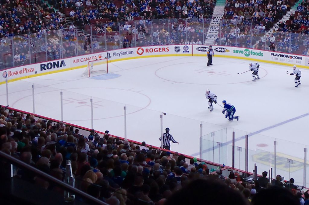 Vier Männer spielen Hockey auf einem Eisstadion, mit zwei in der Nähe des Tors und Zauns stehenden und Zuschauern auf Stühlen im Vorder- und Hintergrund sowie einer Treppe in der Ferne.
