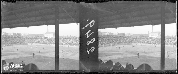 Ein Schwarz-Weiß-Foto eines Fußballspiels in einem Stadion, mit Spielern auf dem Rasen und Zuschauern in den Rängen, vor Gebäuden, Bäumen und einem klaren Himmel, mit dem Text "Piazza della Repubblica, Rom, Italien" in der unteren linken Ecke.