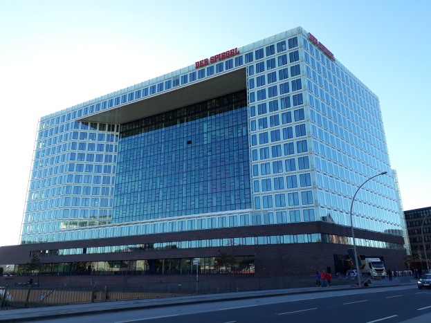 Großes Gebäude mit vielen Fenstern, als Deutsche Bank Hauptquartier identifiziert, umgeben von Straßenlaternen, Straßenlaternen, Kraftfahrzeugen, Fußgängern, Bäumen und einem bewölkten Himmel.