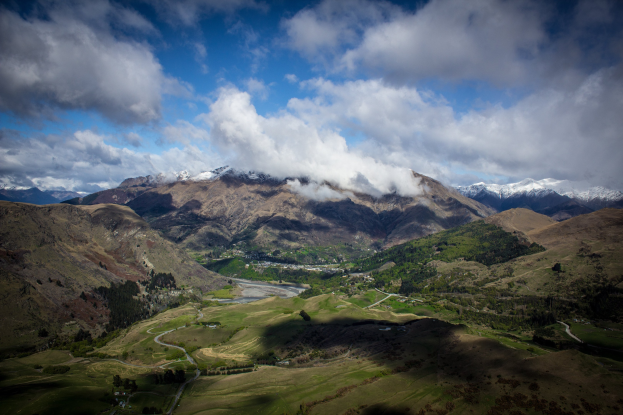 Ein atemberaubender Blick von der Spitze eines Berges in Queenstown, Neuseeland, der üppiges grünes Gras, Bäume, eine gewundene Straße und einen Himmel voller weißer, flauschiger Wolken zeigt.