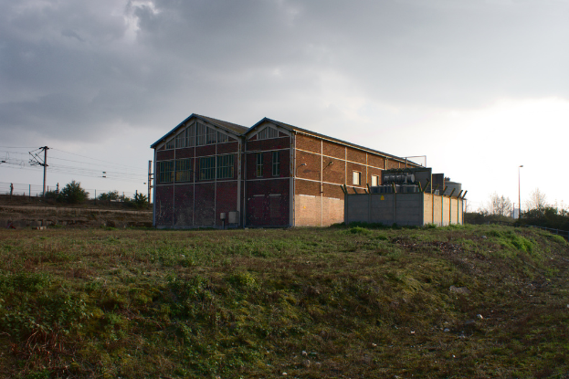 Eine Szene mit Gartenhütten, Transformatoren, Strommasten mit Kabeln, Zäunen, einem Straßenmast mit Straßenlaterne, Bäumen und einem bewölkten Himmel.
