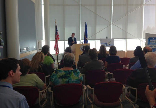 Ein Mann steht an einem Podium und spricht zu einem Publikum, das auf Stühlen sitzt, mit zwei Flaggen neben ihm und einer Wand, Säulen und Fenstern im Hintergrund.