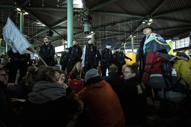 Eine Gruppe von Menschen steht vor einer Menschenmenge in einem Bahnhof, einige halten Schilder und Banner, mit Säulen und Deckenlampen im Hintergrund.