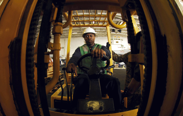 Ein Mann in einer grünen Jacke und einem weißen Helm bedient einen Gabelstapler in einer Fabrik, wobei andere Personen im Hintergrund zu sehen sind.