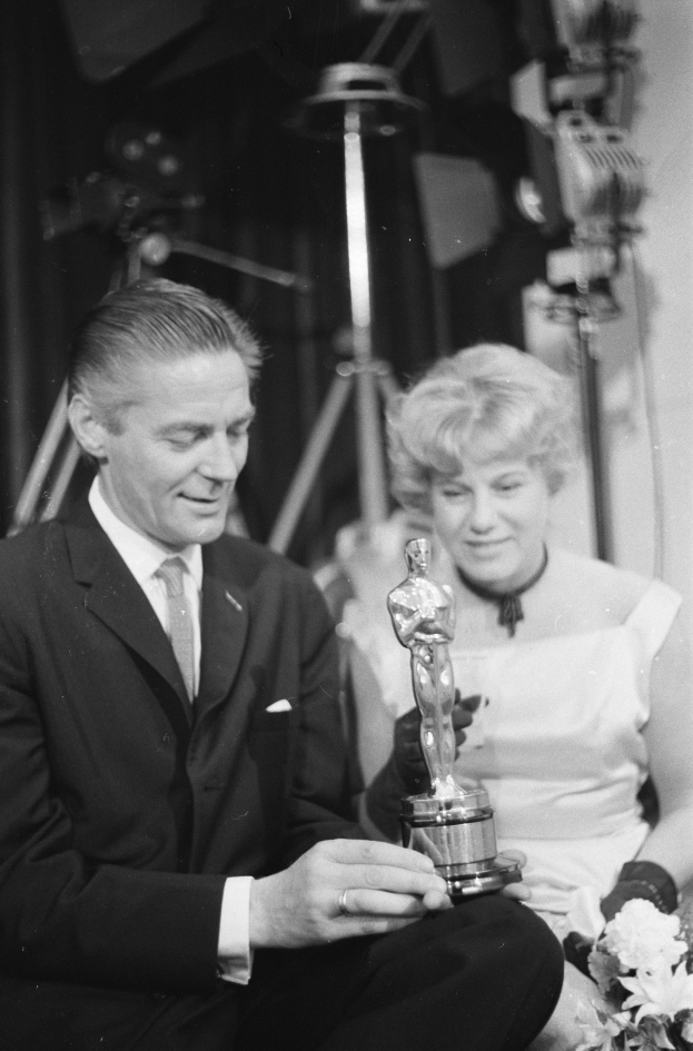Ein Mann in einem schwarzen Blazer, weißem Hemd und Krawatte hält eine Oscar-Statue in der Hand und sitzt neben einer Frau, die einen Blumenstrauß hält, mit Lichtern im Hintergrund, alles in Schwarz-Weiß dargestellt.