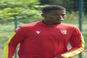 Ein junger Mann in einem roten und gelben T-Shirt steht auf einem Fußballfeld umgeben von einem Zaun und Bäumen und sieht entschlossen aus.