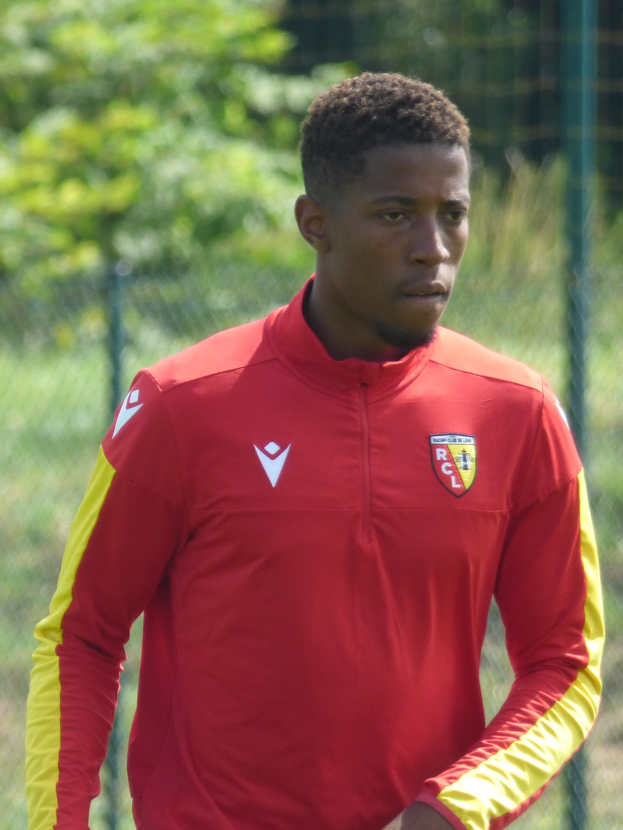 Ein junger Mann in einem roten und gelben T-Shirt steht auf einem Fußballfeld umgeben von einem Zaun und Bäumen und sieht entschlossen aus.