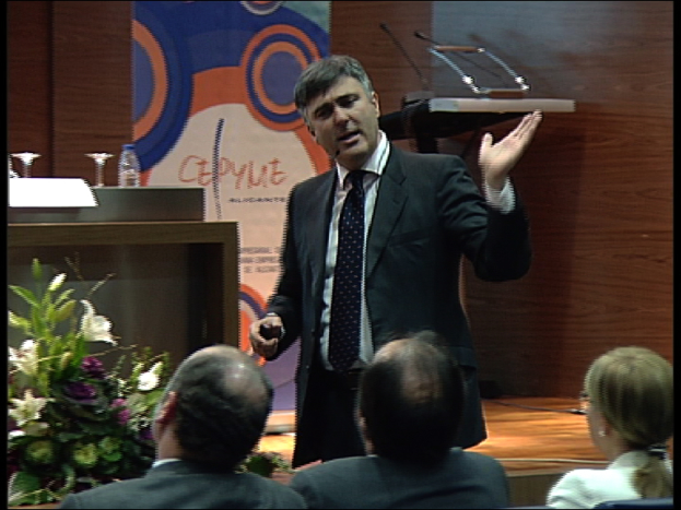 Ein Mann in einem schwarzen Blazer steht an einem Podium mit Mikrofonen, spricht in ein Handmikrofon, mit einer Blumenanordnung und einem Tisch mit verschiedenen Gegenständen daneben, während drei Personen in Stühlen vor ihm sitzen, vor einer braunen Wand und einer bunten Tafel mit Text.