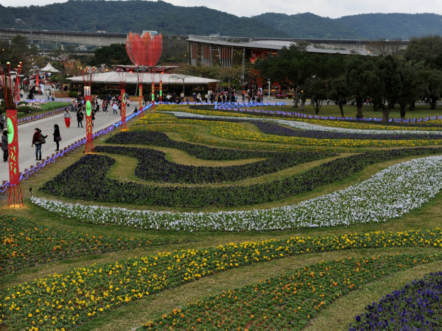 Ein großer Blumen Garten in einem Park mit Menschen, die herumlaufen, umgeben von grünem Gras, Bäumen, Gebäuden, einer Brücke, Bergen und einem klaren blauen Himmel, mit Pfählen und Lichtern auf der linken Seite.
