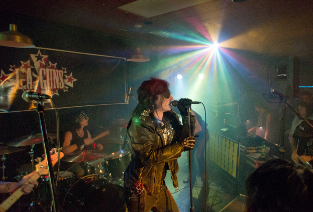 Eine Frau in einer schwarzen Jacke singt in ein Mikrofon auf der Bühne, mit einem Mann in Sonnenbrille und Stirnbinde, der ein Instrument spielt, und einem Plakat mit der Aufschrift "LA Guns" im Hintergrund, unter Diskoleuchten und hängenden Lichtern.